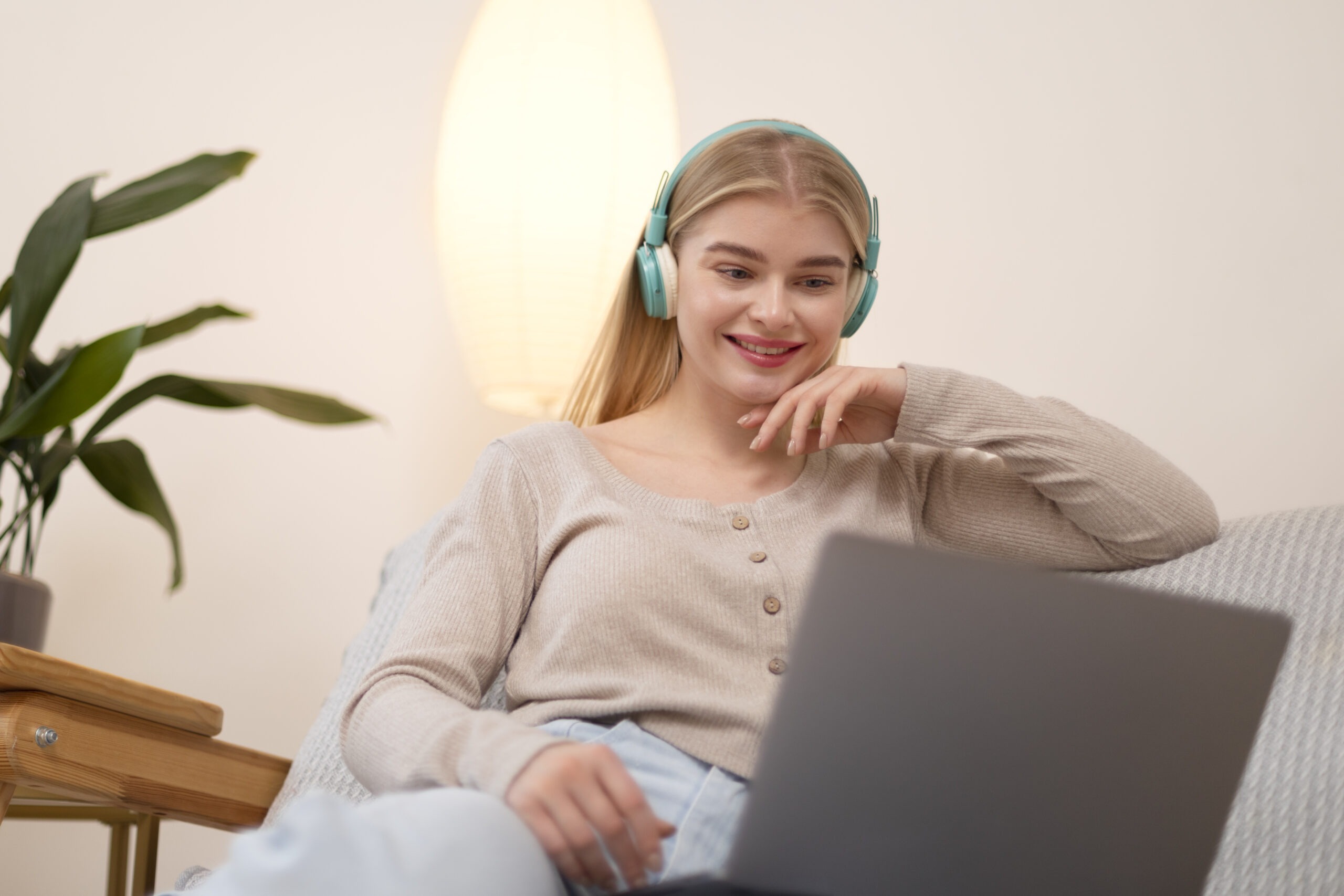 Woman Wearing Teal Headphones Smiling While Using a Laptop on a Cozy Couch